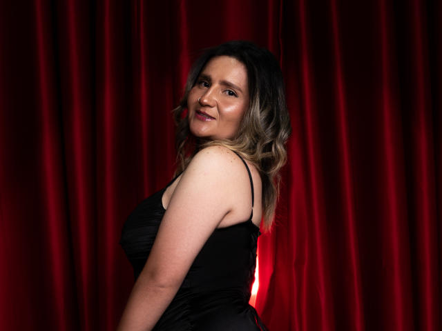 LiliEvanss with wavy blonde hair, smiling, wearing a black dress, showing her boobs and ass, standing against red curtains. She's cute and has a sexy, confident look.