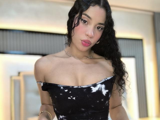 LunaVali with curly black hair, pink lipstick, and a black off-shoulder dress with white patterns, wearing a pearl necklace, looking at the camera, showing her small tits.