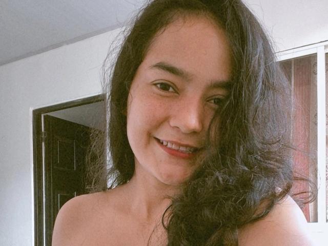 LilianaPink has wavy black hair, brown eyes, and a cute smile with visible teeth. She's topless, showing her bare shoulders. She's standing in a room with a door.