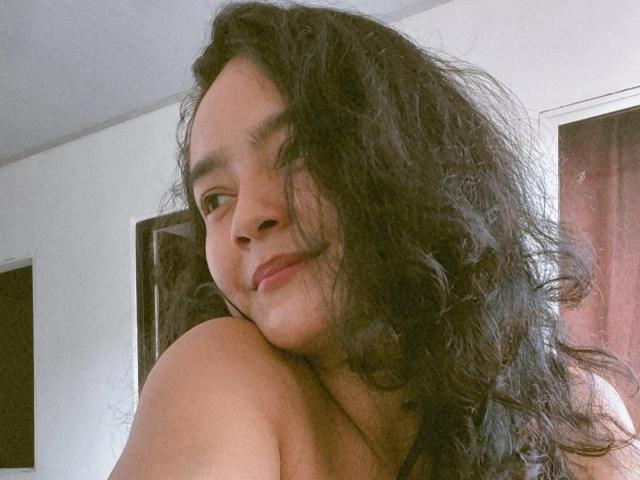 LilianaPink with wavy black hair, smiling softly, showing bare shoulders, her small tits slightly visible, in a casual, natural light setting.
