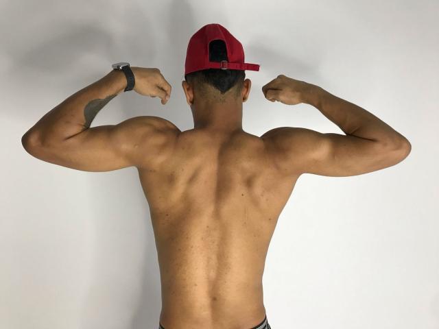 HonyAndMcKensy with muscular back, arms flexed, wearing a red backward cap, black wristband, and black underwear, showcasing a tattoo on the right arm.