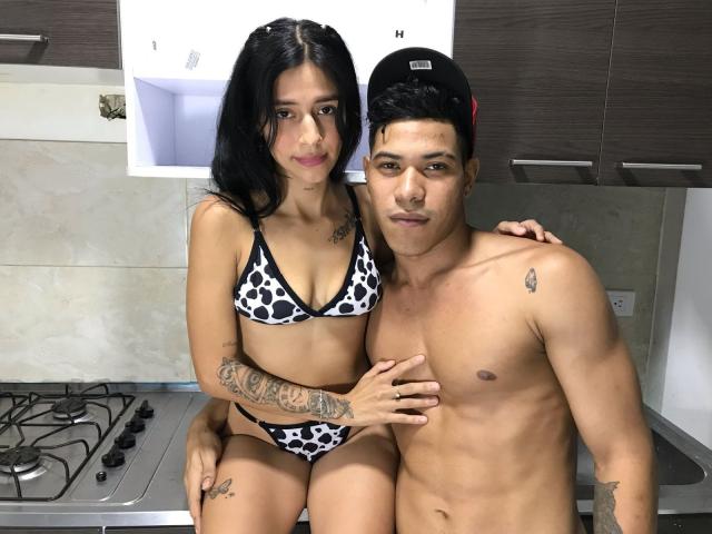 HonyAndMcKensy with black hair in a cowprint bikini, sitting on a shirtless McKensy with tattoos, in a kitchen. McKensy's hand on HonyAndMcKensy's hip, wearing a black cap.