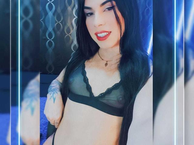 VinnaSkyHott with long brown hair, blue bra, tongue out, middle finger up, showing her large tits, pale skin, and a necklace, taken from a low angle.