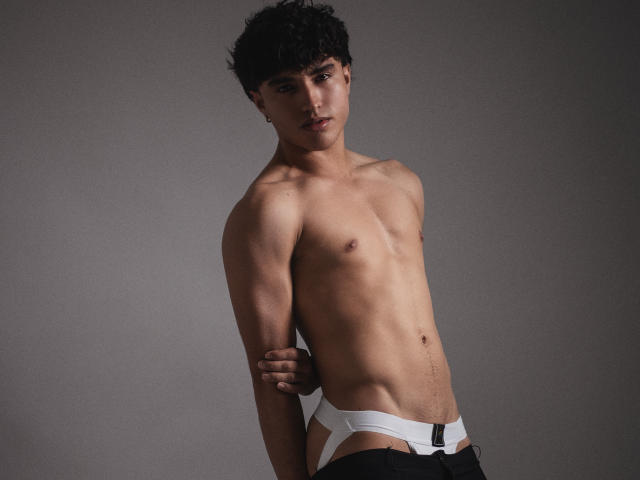 ZaneCross, shirtless, with dark curly hair, black short-sleeve shirt, hand on chin, sitting, looking serious, exposing a hint of his bare ass, against a neutral gray background.