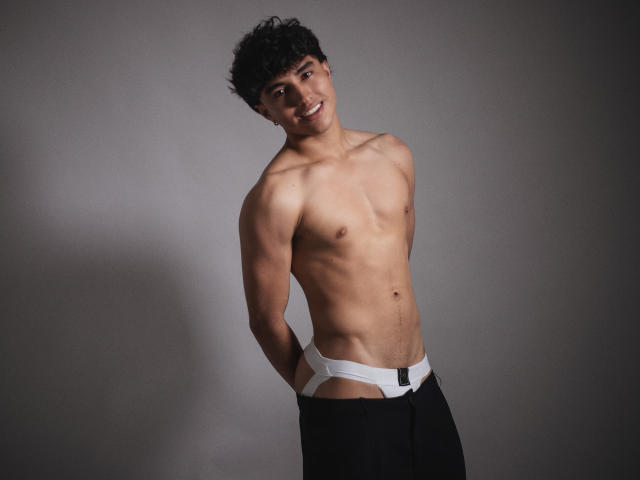 ZaneCross has a lean, muscular build, wearing black pants with a white waistband, exposing his butt cheeks. He's shirtless with short curly black hair, looking serious.