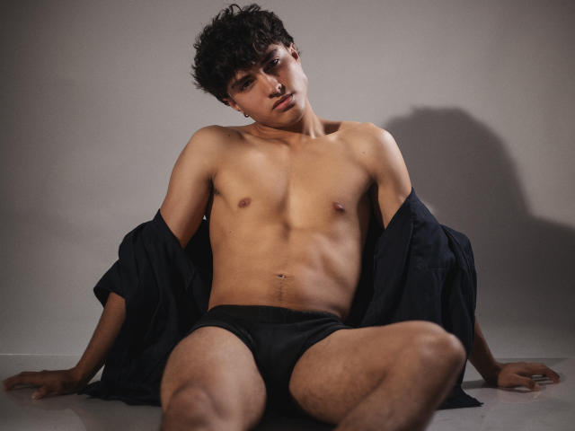 ZaneCross, shirtless, wearing black pants and a white jockstrap, smiling, hands in pockets, short curly black hair, toned, tan skin, standing against a gray background.