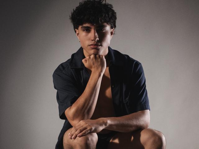 ZaneCross, shirtless with curly dark hair, black underwear, and open dark shirt, sitting with legs apart, revealing a slim, toned body and small, flat ass.