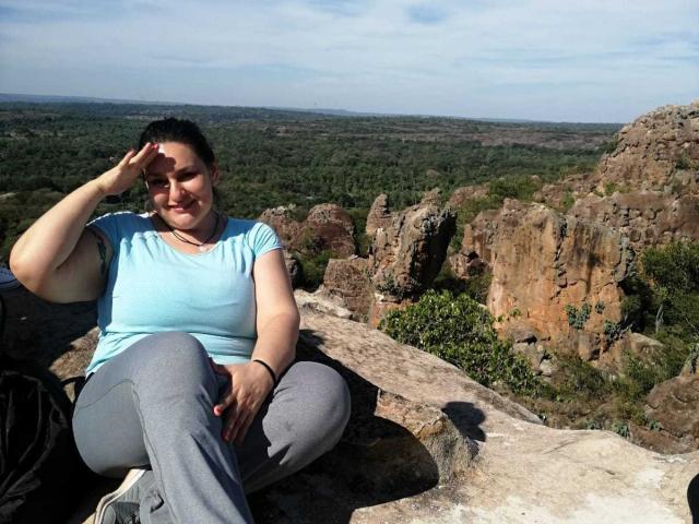 LadySelene, chubby with small tits, wearing a blue shirt and gray pants, sits on rocky terrain, smiling, hand on forehead, in a sunlit, rocky landscape with green trees.