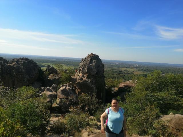 LadySelene stands with her tits in a light blue shirt, dark hair, and sunglasses, in front of rocky terrain and green bushes under a bright blue sky.
