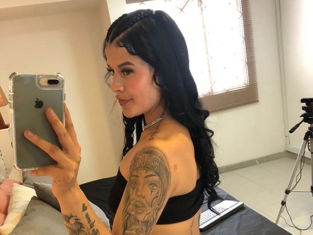 Luciana Dartois con reggiseno nero, capelli lunghi e ricci, treccia frontale, tatuaggi sul braccio destro, che tiene un telefono, sorride allo specchio, camera da letto, treppiede sullo sfondo, laptop sul letto..