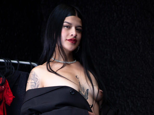 Luciana Dartois, con lunghi capelli neri, rossetto rosso e collana nera, senza top, che mostra tatuaggi e piccoli seni, con una giacca nera tolta, che tiene in mano della lingerie rossa..