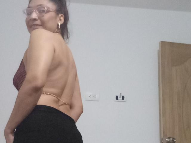 PalomaSexyBeauty with glasses, tan skin, dark hair in a ponytail, topless with back to camera, showing small gold chain, black pants, and smiling. Her tits are visible from side.