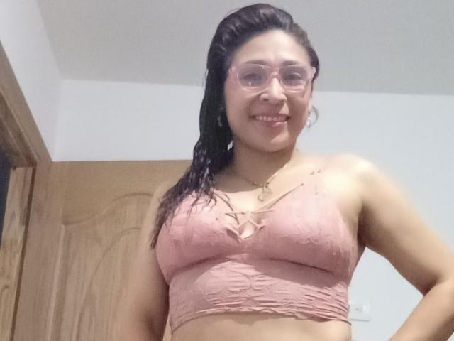 PalomaSexyBeauty with wet brown hair, glasses, and pink lace bra, smiling, showing her tits, standing in front of a wooden door.