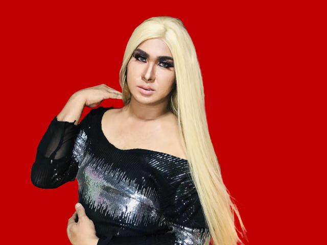 BornStarTS with long blonde hair, black sequined top, dark makeup, and a hand touching her hair, against a red background. Boobs are visible under the top.
