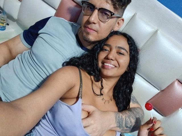 LeoAndKali with curly black hair, smiling, in a light purple top, holding a red lollipop, while LeoAndKali with curly brown hair, glasses, and a grey shirt, touches her tits.