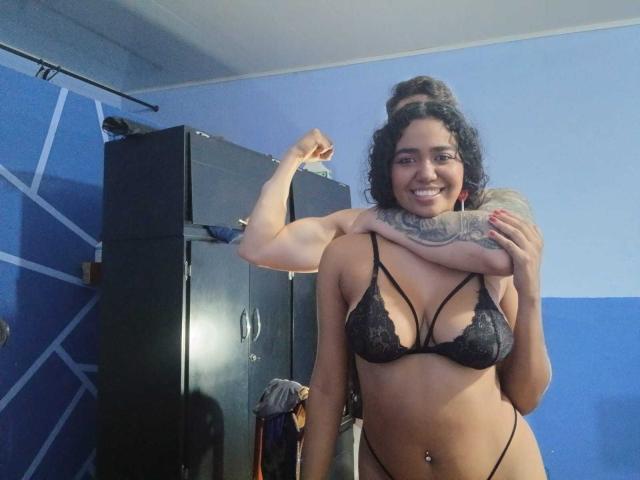 LeoAndKali with curly dark hair, smiling, wearing black lace bra and thong, arm tattoo, flexing bicep, black locker in background, blue wall, belly button piercing.