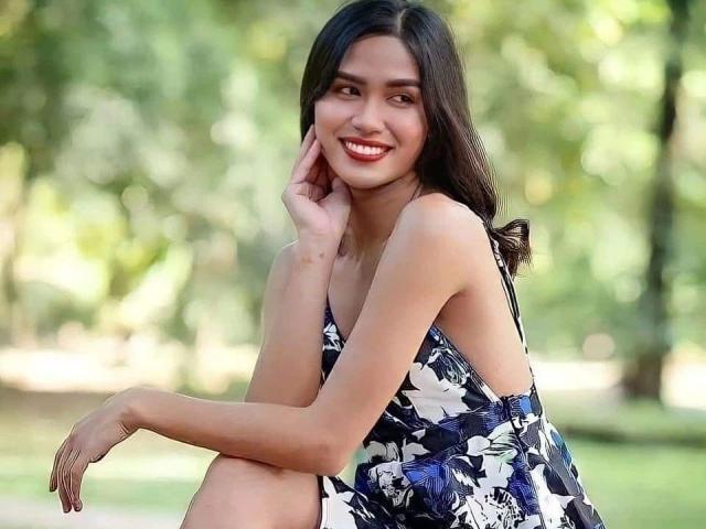 MariaPeters-hot with long dark hair, smiling, wearing a black and white floral dress, sitting, showing her tits, hand on cheek, green blurred background, sunny day, red lipstick.