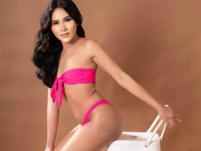 MariaPeters-hot with long black hair, wearing a pink bikini, has a slim, sexy body, small tits, and a cute ass, sitting on a white chair against a brown background.