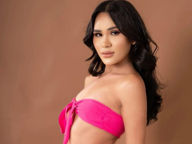 MariaPeters-hot with long dark hair, pink strapless top, tied below her small, perky boobs, light brown skin, subtle makeup, and a neutral expression, against a brown background.