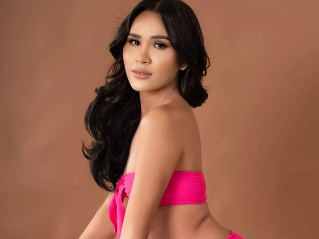 MariaPeters-hot with long black hair, pink strapless top, and medium tits, looking at camera with a slight smile. Soft brown background. Her ass is partially visible.