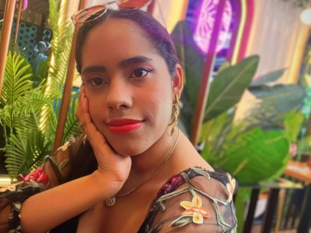 Sienna Taylor, con rossetto rosso, la testa appoggiata sulla mano, indossa una blusa trasparente con fiori, orecchini dorati e occhiali da sole in testa, sorridendo dolcemente. La sua pelle scura e i capelli neri sono visibili..