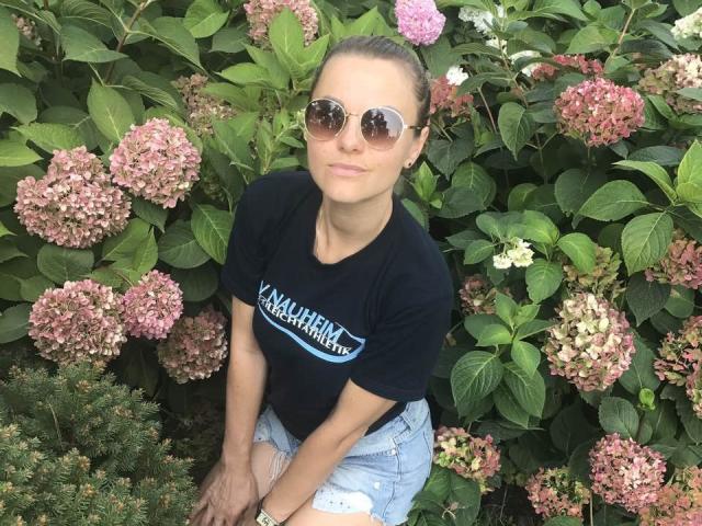 BelleLisaG with brown eyes, tied-back brown hair, wearing round sunglasses, black t-shirt, and ripped denim shorts, standing in front of pink and white hydrangeas.