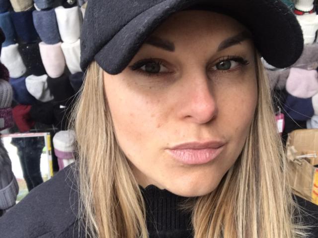 BelleLisaG has blonde hair, a black cap, and thick eyebrows. She's wearing a black turtleneck with her pink lips slightly pursed. Her green eyes are looking at the camera.