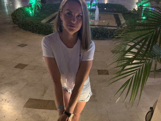 BelleLisaG with blonde hair, white t-shirt, and denim shorts, standing and leaning forward, hands on knees, smiling slightly, green wristband, palm leaves to the right.
