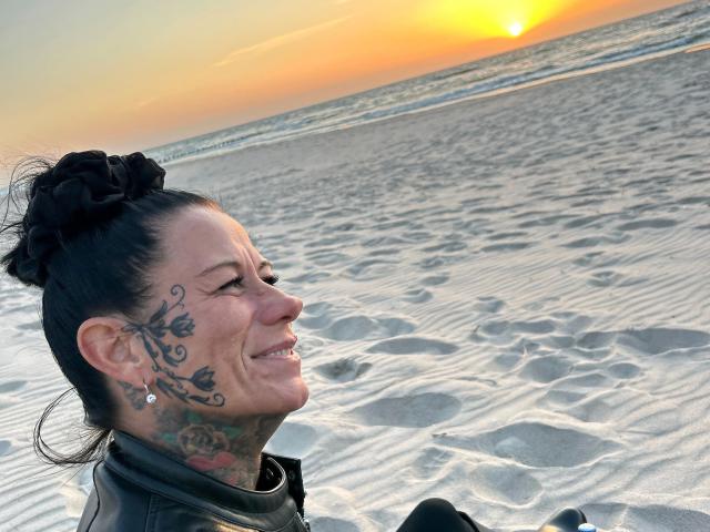 LucyLove-hot with black hair in a bun, smiling, showing tattoos on her face and neck, wearing a leather jacket, at a sandy beach during sunset.
