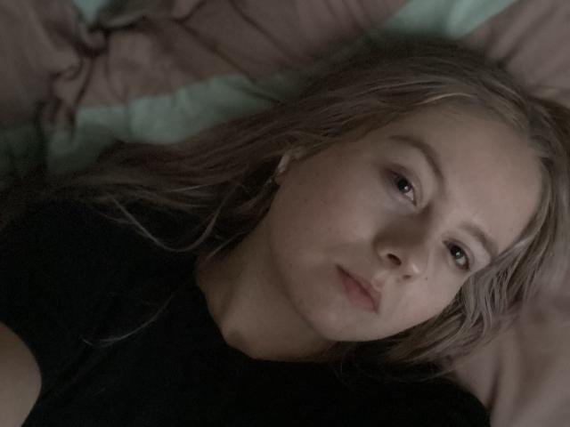 KittyAimee heeft lichtblond haar, een bleke huidskleur en ligt op een bed met een zwart shirt, met een lichtjes fronsende uitdrukking en een subtiele, ontspannen gezichtsuitdrukking..