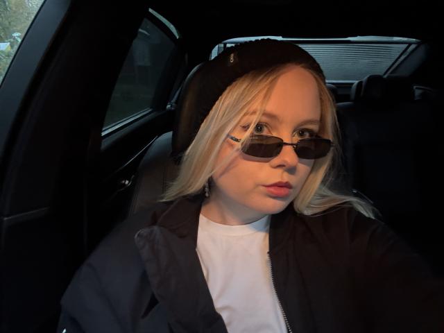 KittyAimee a les cheveux blonds, porte une casquette noire, des lunettes de soleil noires, une chemise blanche, une veste noire, et a les lèvres rouges. Elle est dans une voiture et regarde directement l'appareil photo..