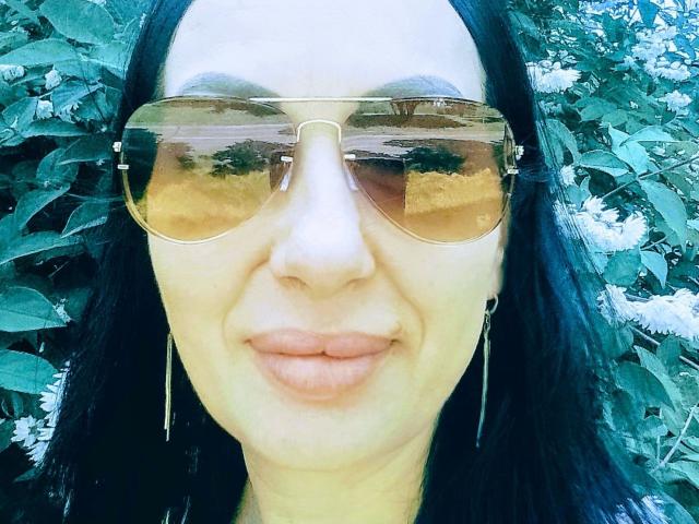 MirandaHott69 with tan aviator sunglasses, black hair, and pink lips, wearing long earrings, smiling slightly, reflecting trees in her sunglasses.