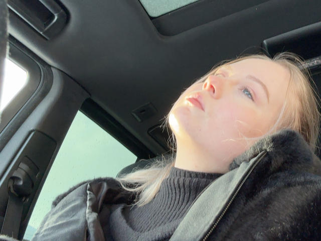 KittyAimee tiene la piel pálida, ojos azules y cabello rubio, y lleva un suéter negro de cuello alto y una chaqueta con forro de piel, sentada en un coche con la luz del sol en su rostro..