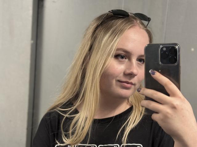 KittyAimee, con cabello rubio, una camiseta negra y gafas de sol en la cabeza, está tomando una selfie con un iPhone negro. Tiene la piel clara y una sonrisa sutil..