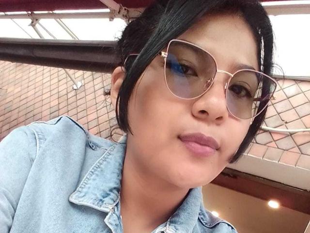 ScarletKry with medium brown skin, black hair, wearing glasses, and a denim shirt, has a slight smile, and her ass is partially visible in the background.