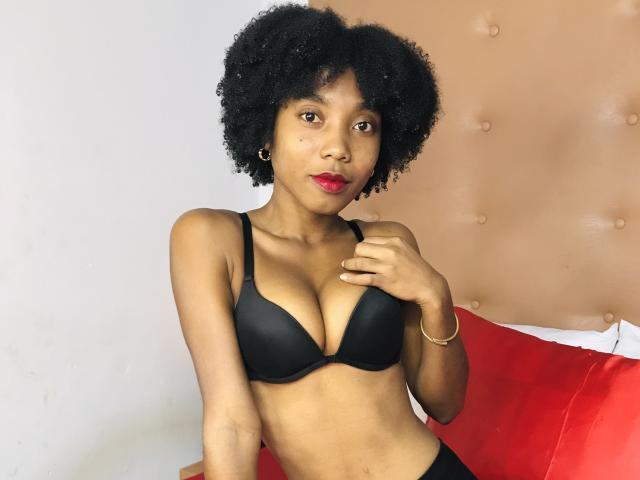TynnaMagik with curly black hair, wearing a black bra, showing her small tits, red lips, gold earrings, and a gold bracelet, sitting on a bed with red pillows.