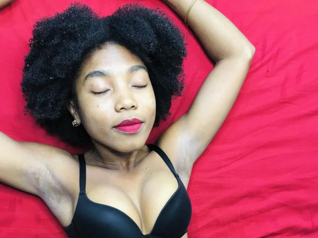 TynnaMagik with dark curly hair, eyes closed, red lipstick, black bra, arms above head, lying on red bed, small earrings, smooth dark skin, small nose, small breasts, relaxed.