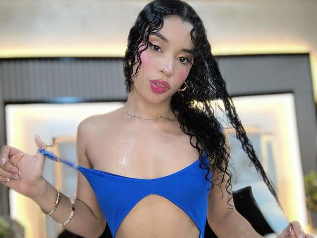 LunaVali with curly black hair, wearing a blue one-piece with cutout sides, showing her tan skin, small belly button piercing, and holding the top of the outfit.