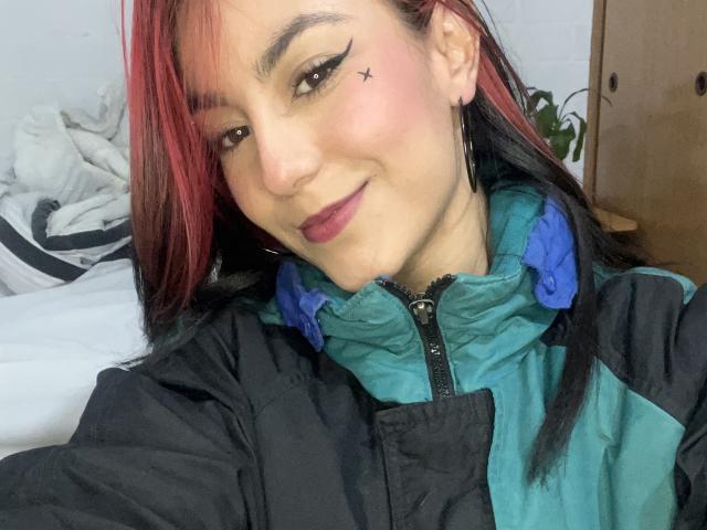 Il viso di AmyStreetwise69, con rossetto rosso, capelli castano con punte rosse e una mano sul viso. Ha lunghe ciglia e un leggero sorriso. La sua pelle è liscia e indossa una maglietta nera..