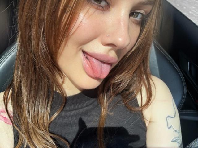 EmmaaRodriguez with black hair, wearing a red and black checkered shirt, leaning on a black car in a forest, showing her tits and ass, with a finger near her mouth.