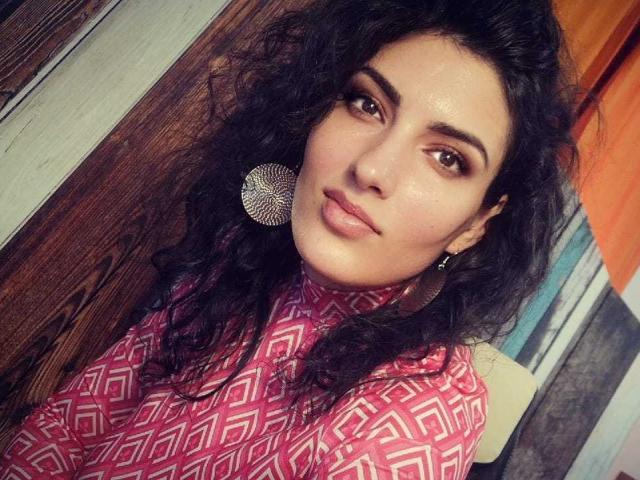 AddictedToUTS with pouty lips, wearing pink geometric pattern shirt, large circular earrings, dark wavy hair, and brown eyebrows, looking to the side with a slightly annoyed expression.