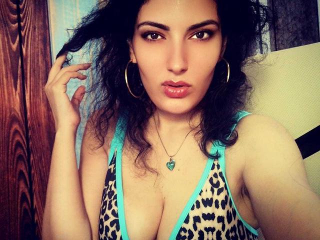 A AddictedToUTS tem cabelo preto encaracolado, olhos castanhos, e usa brincos de prata redondos com uma blusa laranja e rosa, com a mão no cabelo, lábios rosados e finos, em um selfie..