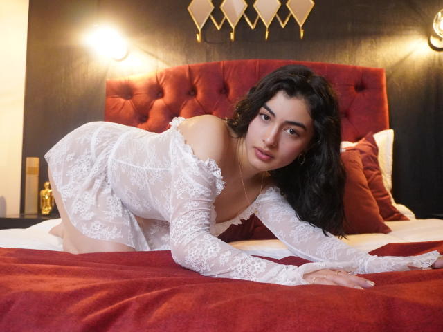 SeleneRoussel, liggend op een bed in zwarte kanten lingerie, met een bril op, een afstandsbediening in haar hand, haar billen omhoog, roze onderbroek, een toetsenbord op het bed, een gouden enkelbandje, ontspannen, sexy..