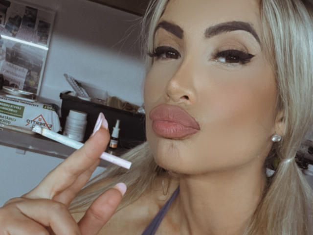 Lellie with blonde pigtails, full lips, and thick eyebrows, holding a makeup brush near her pink-lipped, sexy face, wearing diamond earrings, in a selfie.
