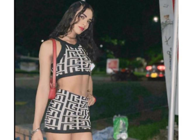 SexyHannaD with long black hair, wearing a black and white geometric crop top and skirt, red backpack, standing at night, red lipstick, tattoo on right side, holding bag.