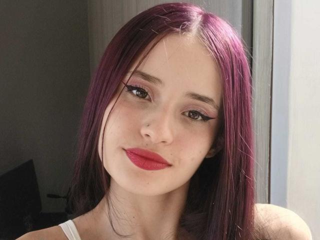 Sweetkim13 with purple hair, red lipstick, black eyeliner, nose ring, and white tank top, smiling at the camera. Her tits are not visible.