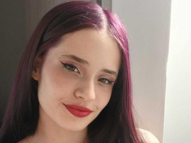Sweetkim13 with purple hair, red lipstick, and a nose piercing, sporting thick black eyeliner and a subtle smile, looking directly at the camera.