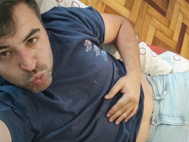 Vio69x's puffy, pink-assed face with dark eyebrows, brown eyes, short black hair, and slight beard, lying on a gray couch in a blue shirt, looking up.