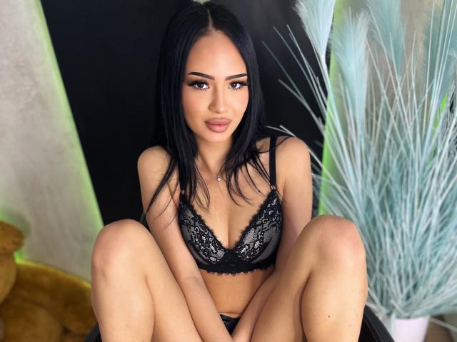 AngelaDestiny with black lace bra, sitting with knees up, showing her tits. She has long black hair, brown eyes, and a cute face. Her pussy is not visible.