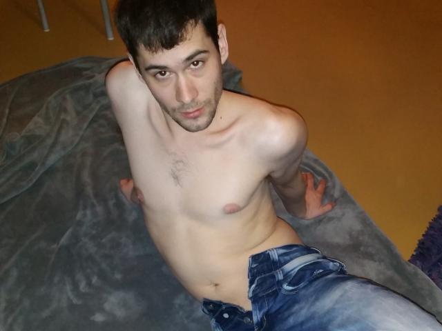 Ethan69, shirtless, lean with small chest, dark hair, looking up, blue jeans, sitting on gray blanket, brown skin, slight stubble, arms behind, casual, relaxed, indoor, slightly mischievous.